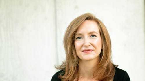 Head shot of Reddy. She wears a black top, light brown (maybe reddish?) hair, against cream backdrop, looks toward camera