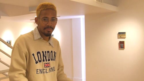 Young, a Black man with glasses & bleached short hair, stands in the white-walled gallery with his sketchbooks.