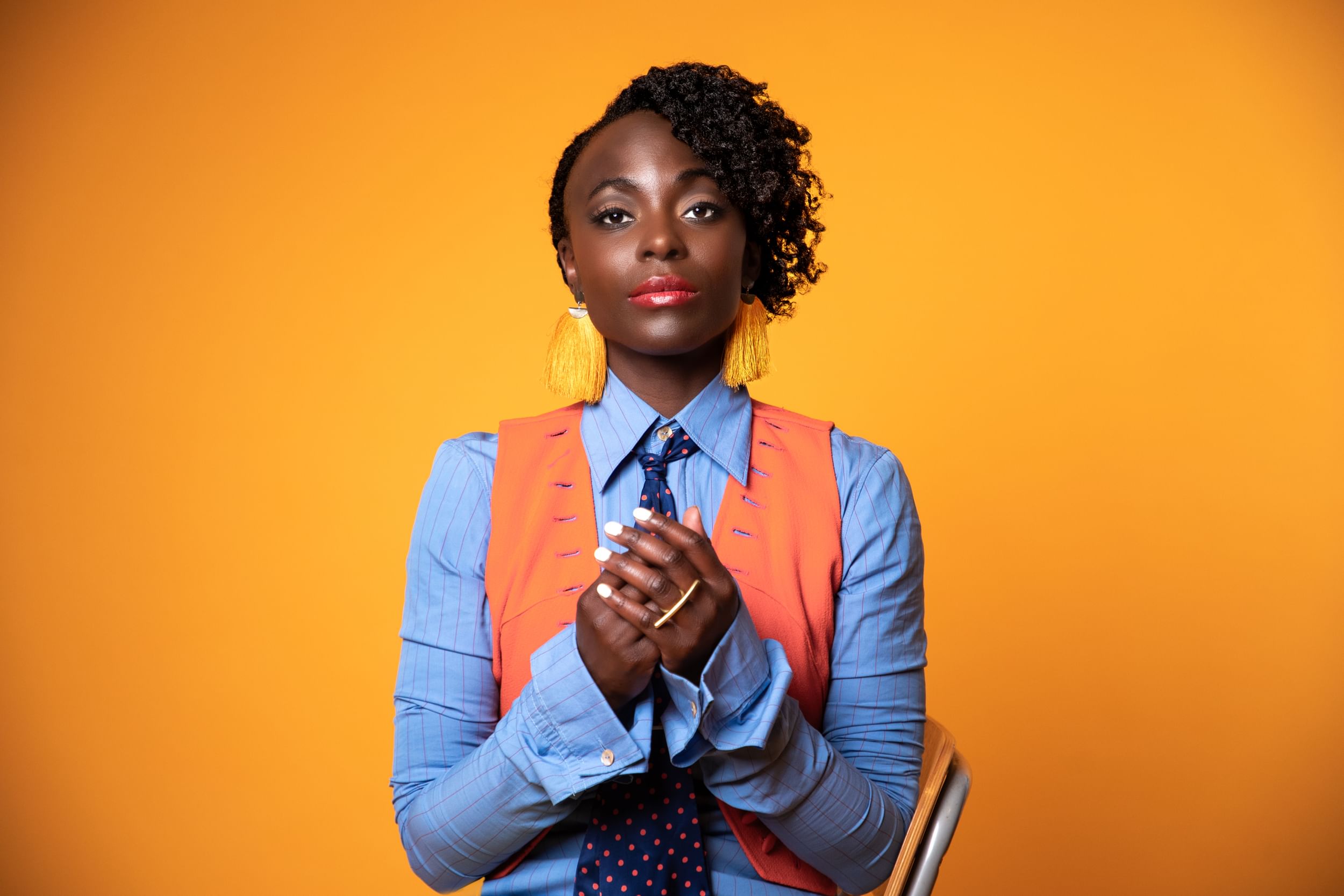 Portrait photo of Joachim, a Black woman, in a blue buttonup w/ tie & vest, hand over hand, over a sharp orange background