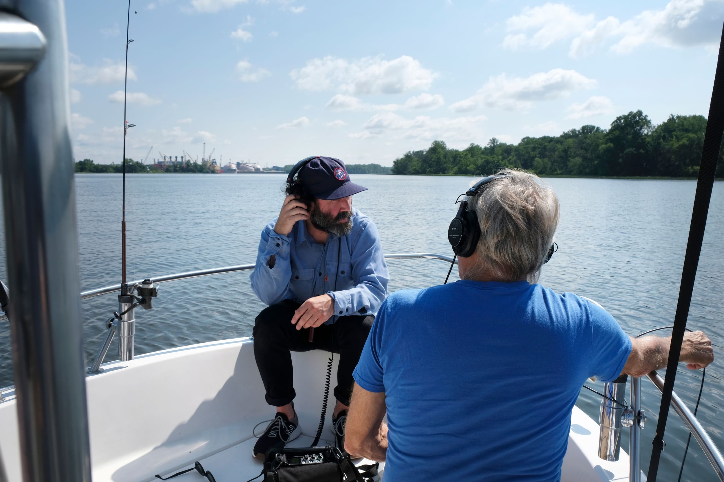 Young, in blue shirt and baseball cap and gray beard, listens to a headset. He sits in the prow of a boat on the calm river.