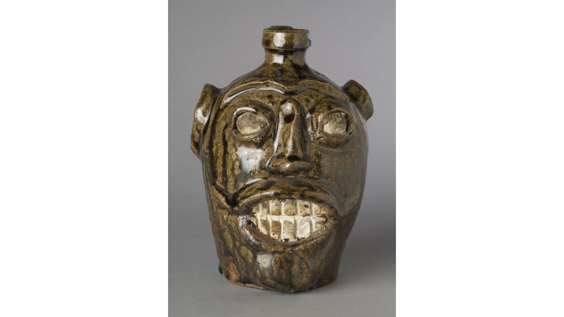 A glazed, earth-toned pottery vessel with a stylized human with large eyes and the front teeth exposed in a grimace or smile.