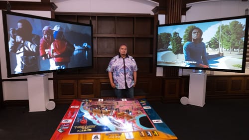 Cabrales, at center, stands between two large TV screens with animated images. A board game about 4 feet square is in front.