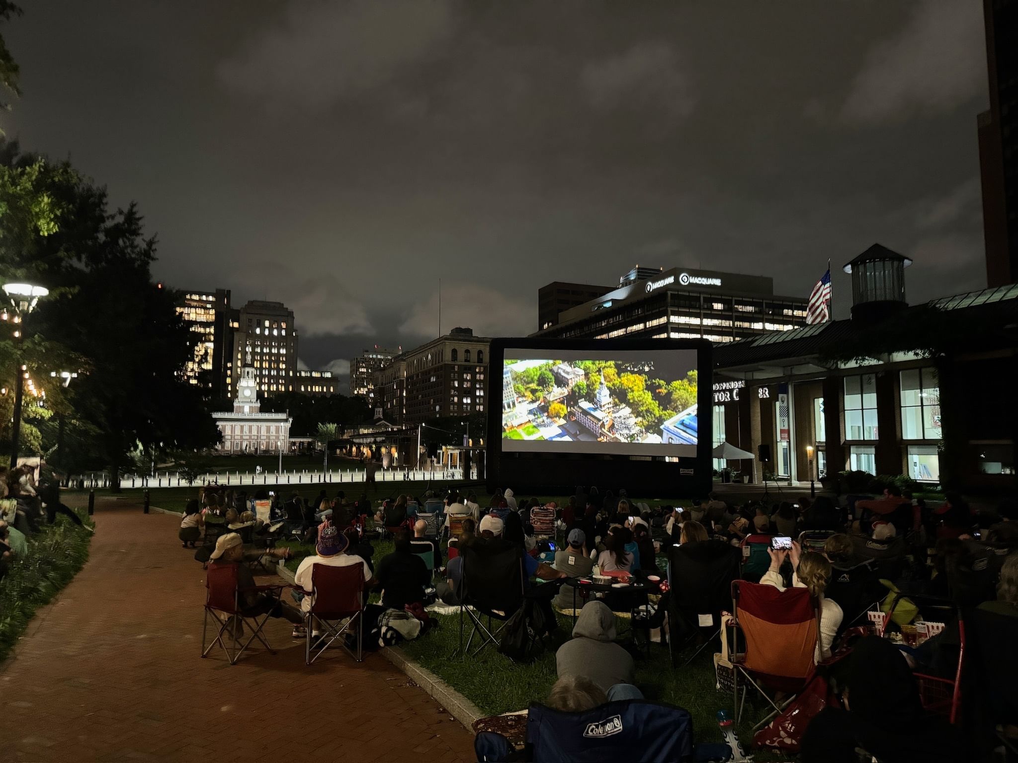 A crowd of people on lawn chairs watch a big outdoor screen on a cloudy summer night beside Independence Hall.