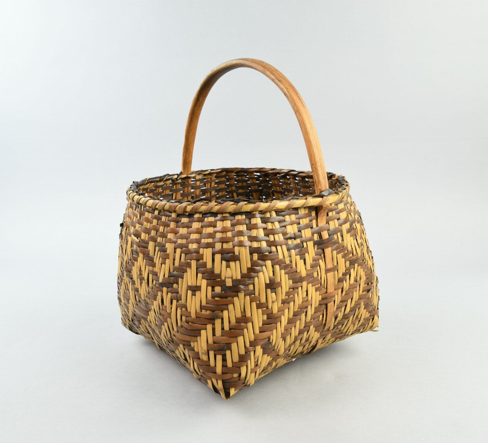 A basket with a curved wooden handle, round top, and square bottom, woven in geometric patterns in yellow and brown.