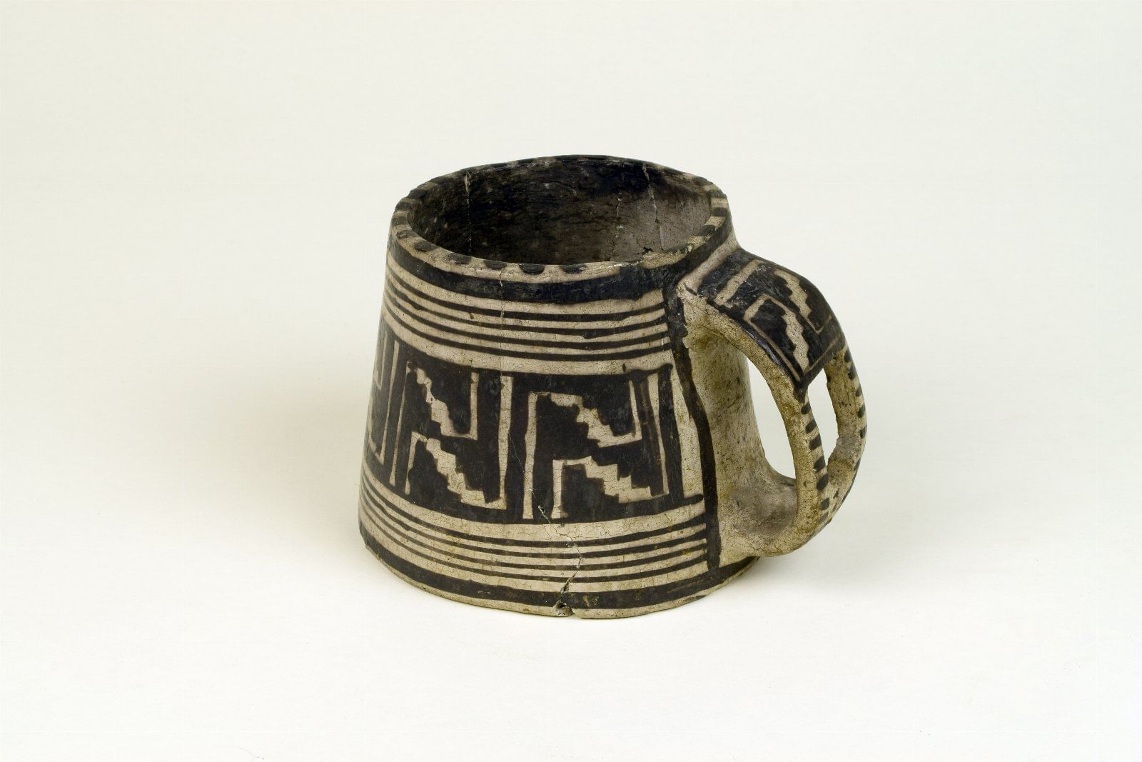 A small authentic clay mug with black-and-white lines and geometric patterns, worn and cracked with age.