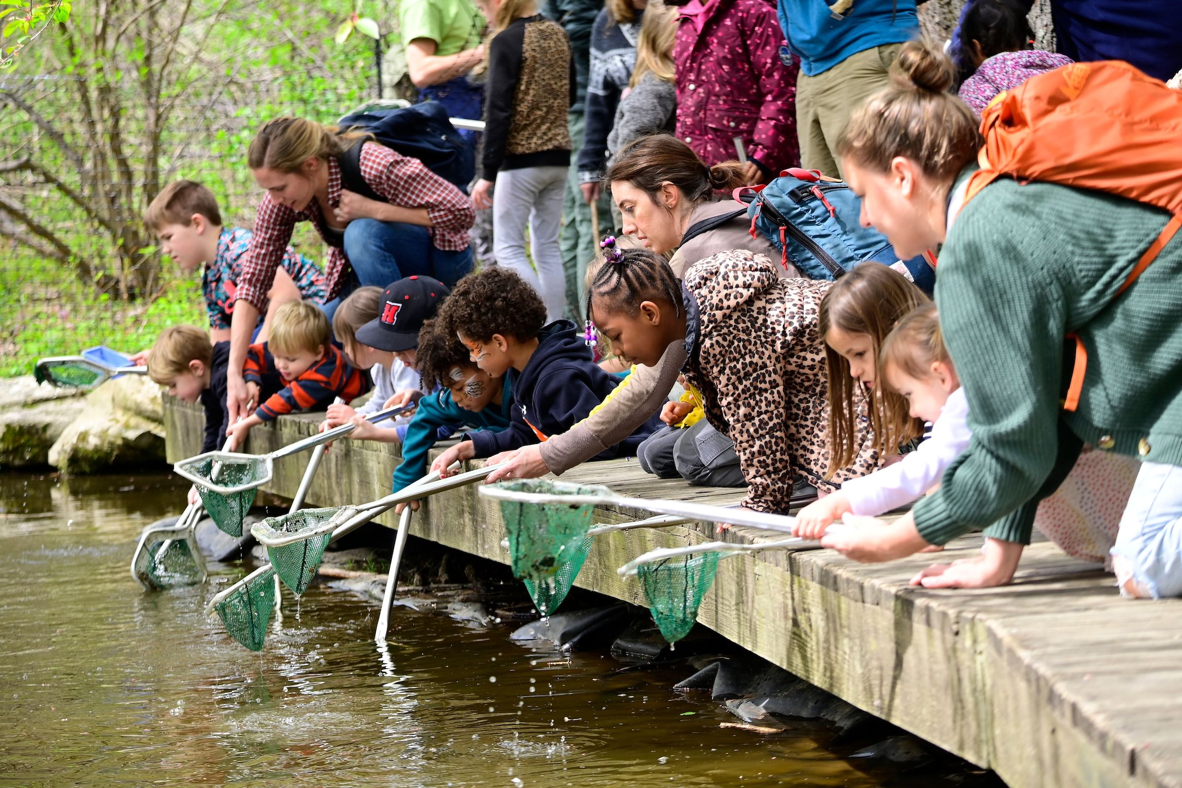A large group of children and adults at the edge of a boardwalk cast nets into a body of water