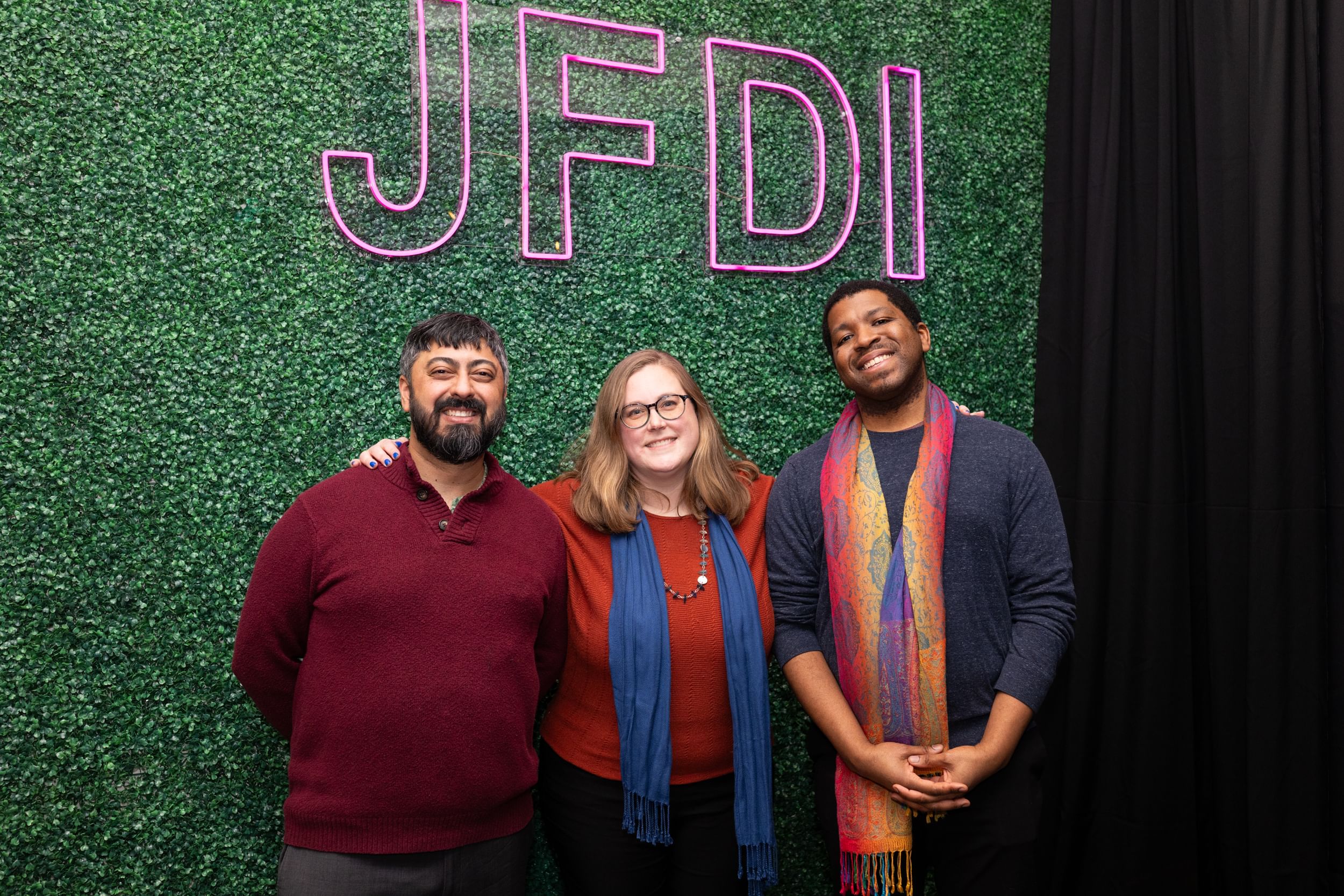 The three BSR leaders stand together smiling in nice sweaters, under pink neon "JFDI" letters.