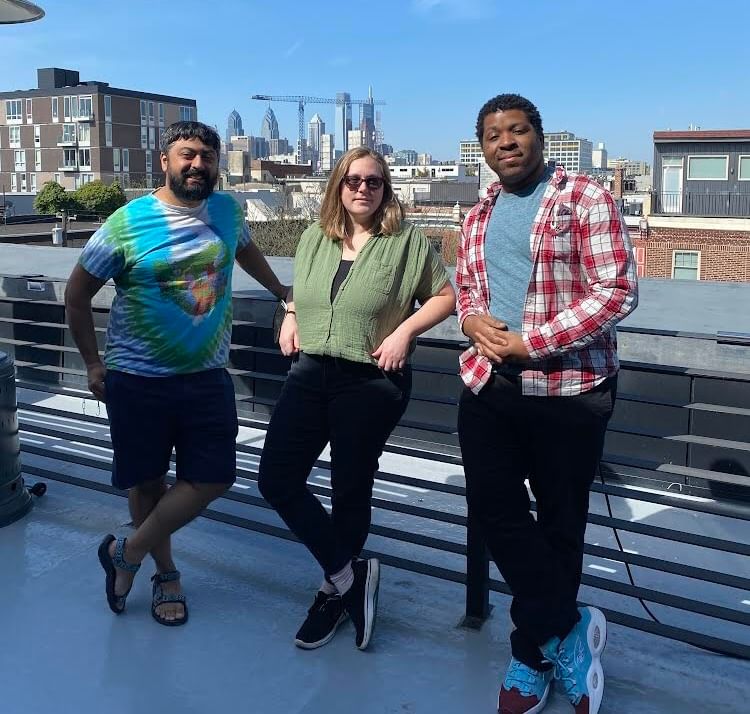 Neil, Alaina, and Kyle lean on the railing of a fourth-floor city deck, smiling in the sun with the Philly skyline behind.