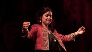 A still of Elkayam mid-performance, her arms out, expressive with her eyes closed. She wears a red studded jacket on stage