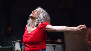 Close-up on Dardaris, a white woman with curly gray hair, standing with arms ecstatically outstretched, in a red blouse.