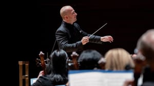 Smiling and wearing a black jacket, Nezet-Seguin conducts, with the heads of a few musicians visible in the foreground.