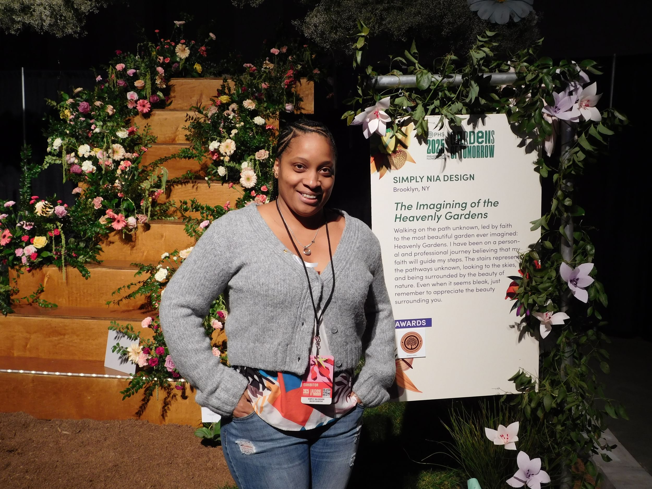 Mozee, a Black woman, poses smiling in a gray cardigan and jeans next to her flower display’s welcome sign.