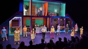 19 smiling Latine people of different ages sing, scattered in front & inside a colorful two-level set like rooms of a house.