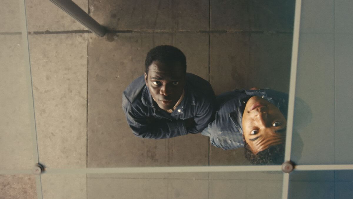 Two young Black men look up at camera, one’s arms are crossed, and seem to be looking through a glass pane.