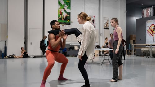 Jackson, a white woman with braided hair, intently watches as Wills, facing Kelly, places a foot on his shoulder.