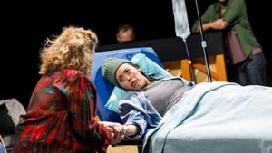 A patient in a hospital bed, head tilted to side to look at someone by their bedside on theater stage
