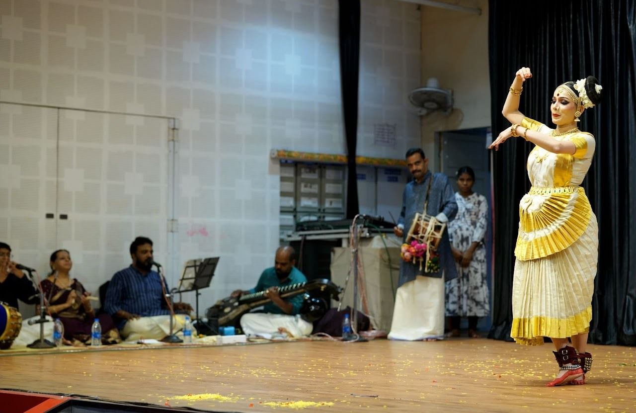An Indian woman in bright yellow and cream colored dress on a brightly lit stage, dancing, with audience watching