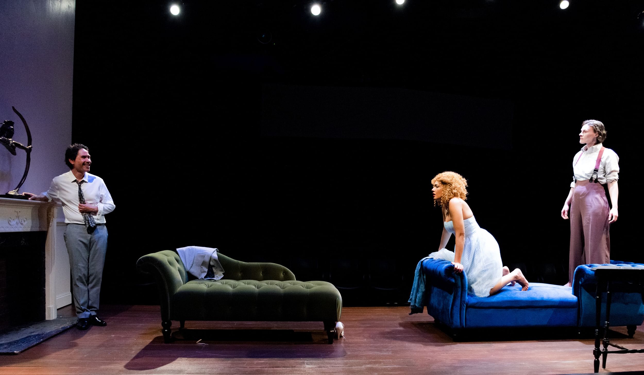 Niebauer & Ladd, on a plush blue chaise, stare in concern at Hernandez, who leans smiling on a fireplace across the room.