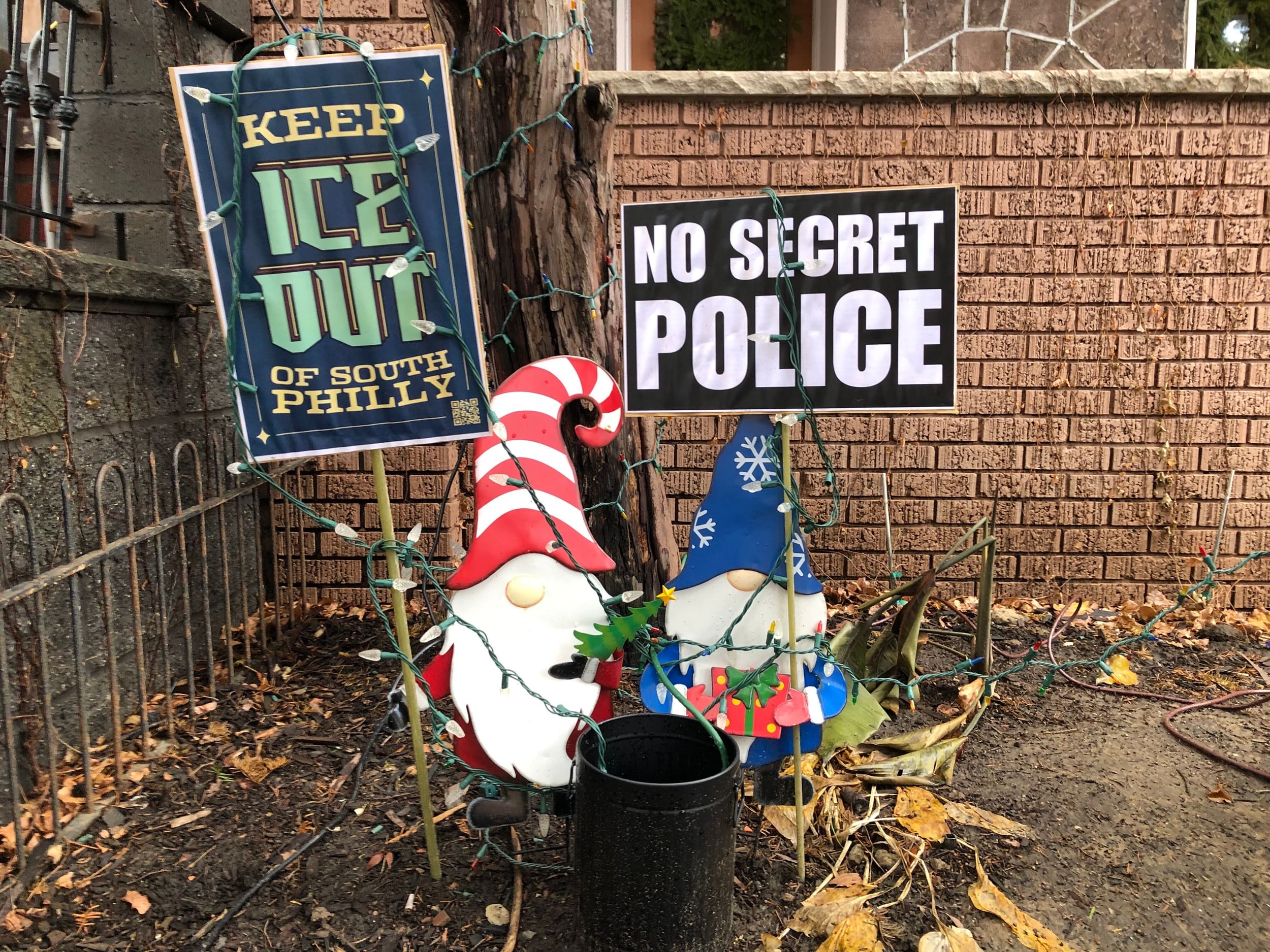 Two Christmas gnomes in a South Philly frontage next to signs that say Keep ICE out of South Philly and NO SECRET POLICE.