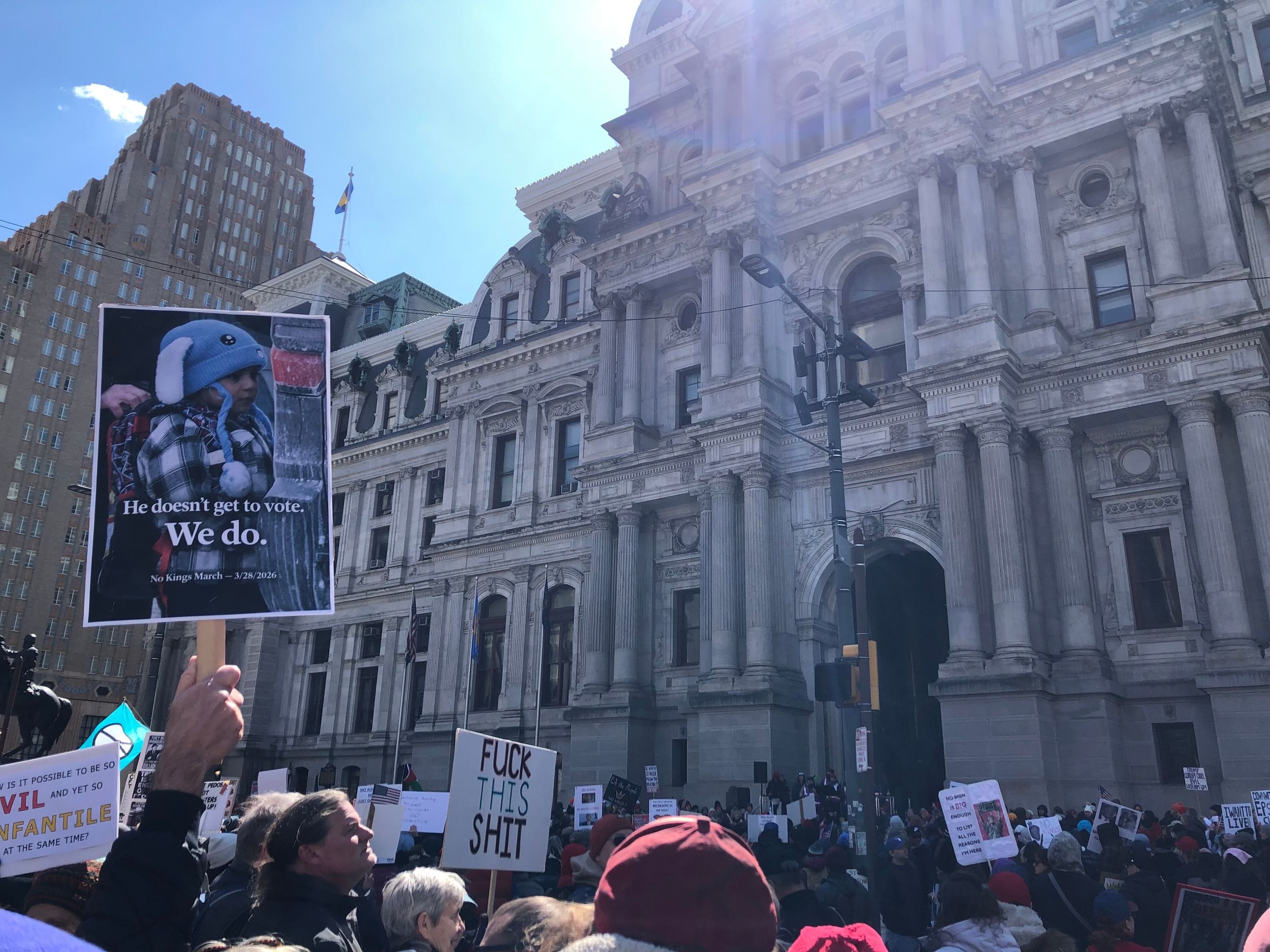 A crowd of hundreds in the shade of the north side of City Hall, with a large sign at left featuring a child detained by ICE.