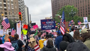 Pic of the crowd of protestors from behind on a cloudy day, with lots of US flags, with a No Kings in America sign at center