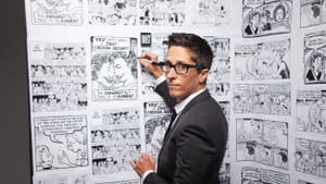 Bechdel in a suit jacket looks over her shoulder at the camera, pencil in hand, comic panels on a wall in front of her.