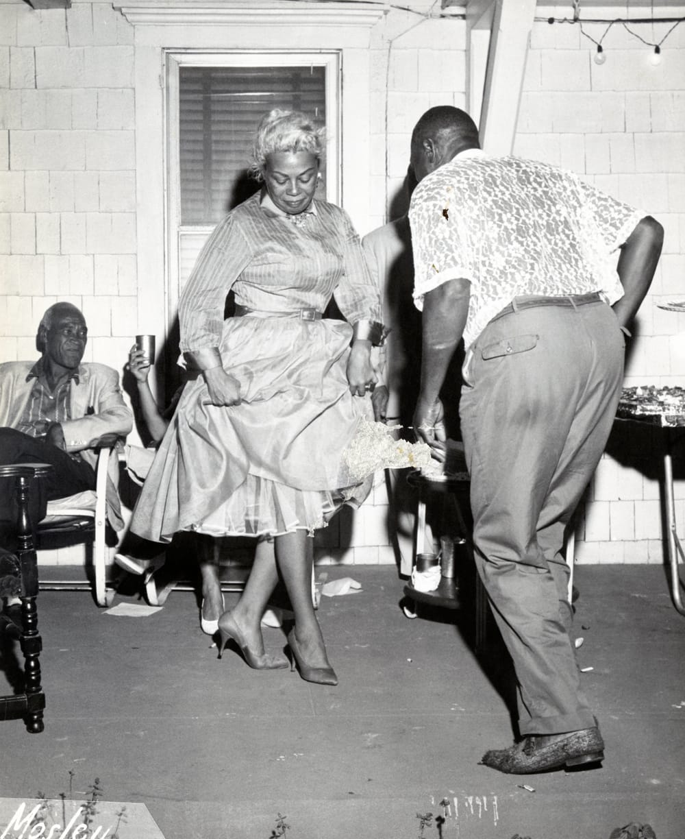 Black and white photo of two older Black people dancing together, in a room with other people talking with each other