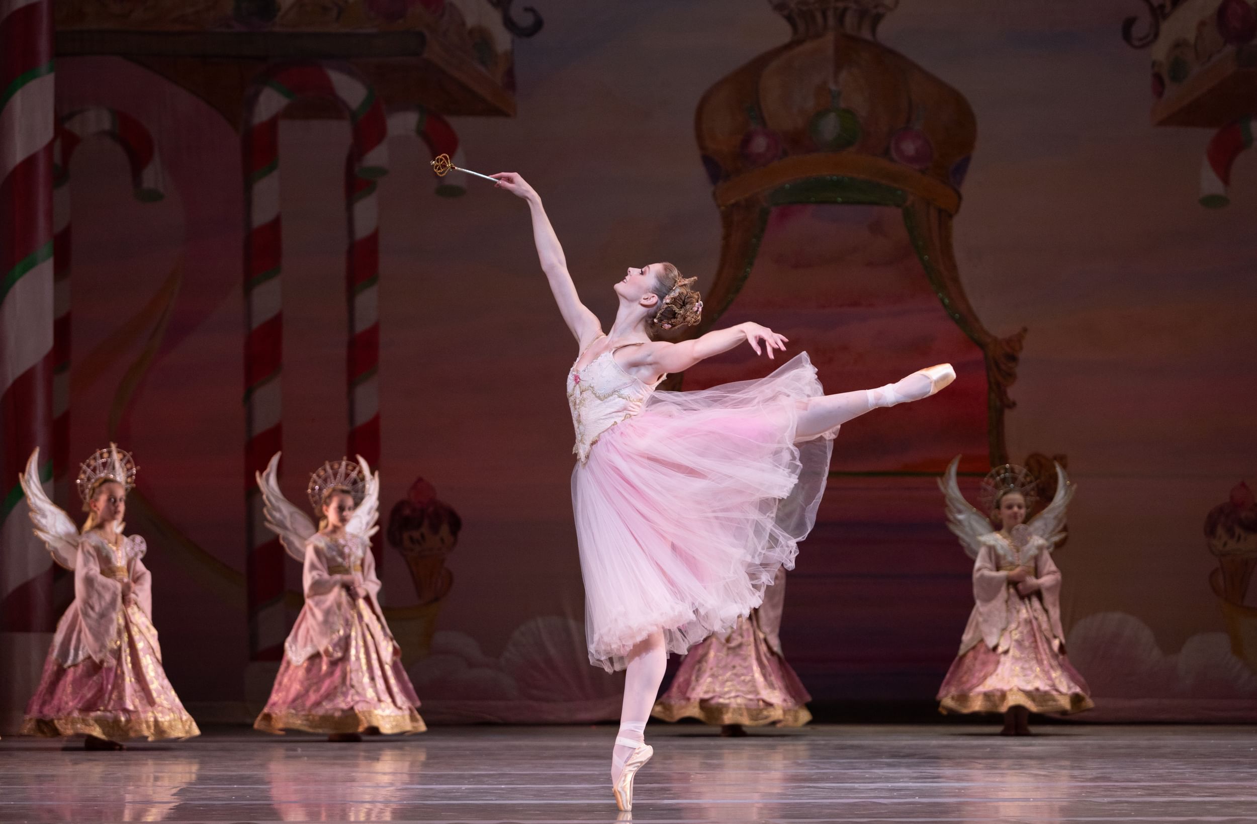 A dancer on stage in white tutu extends her left leg up and out, right arm up, on her right toes. Dancers behind her on stage