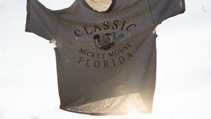 A weathered shirt. Mickey Mouse and ‘Classic Mickey Mouse Florida’ text on it hangs on a clothesline, against a bright sky