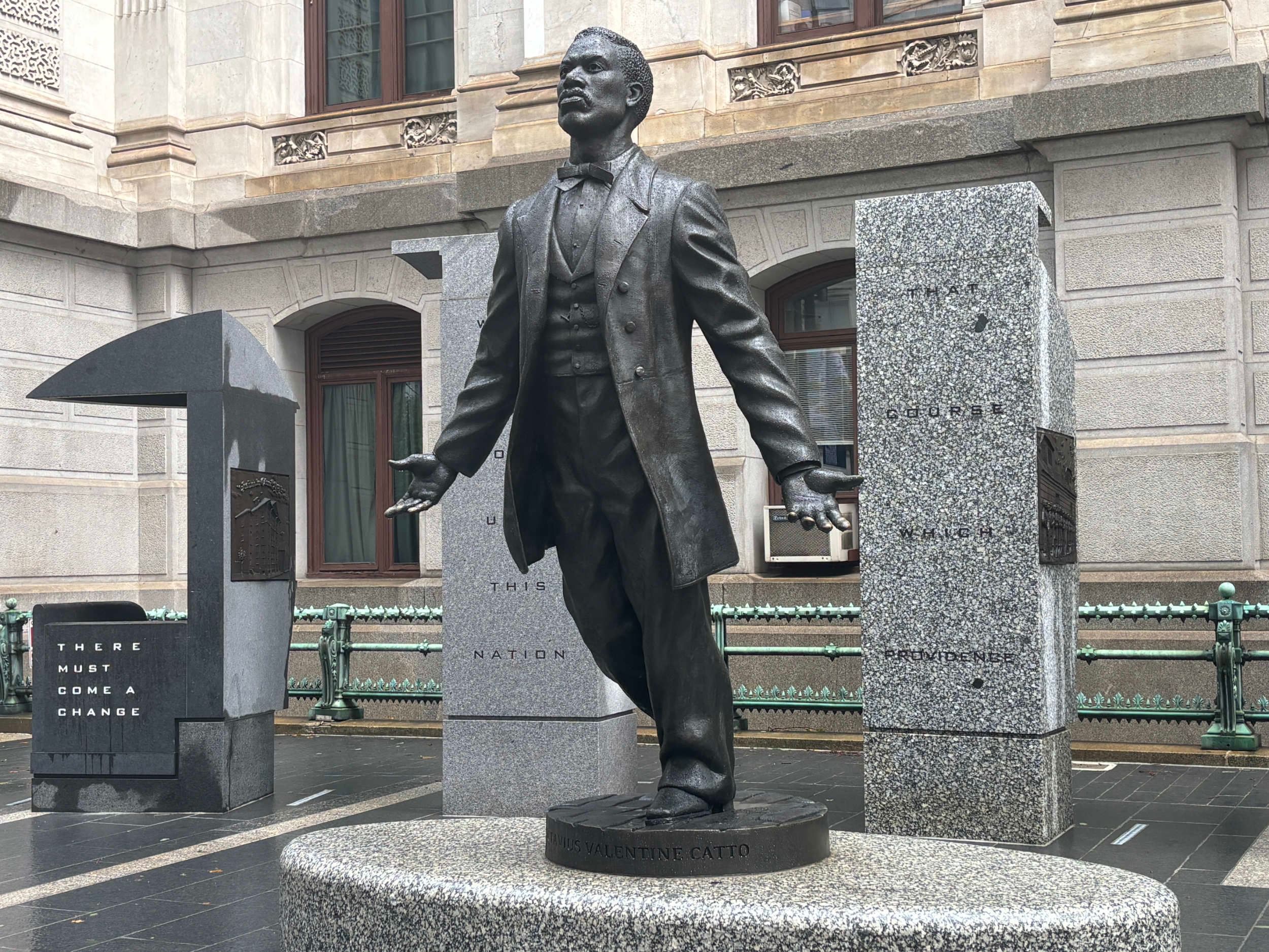 Larger-than-life metal sculpture of Catto, wearing a suit, looking outward, and posing as if about to step off the plinth.