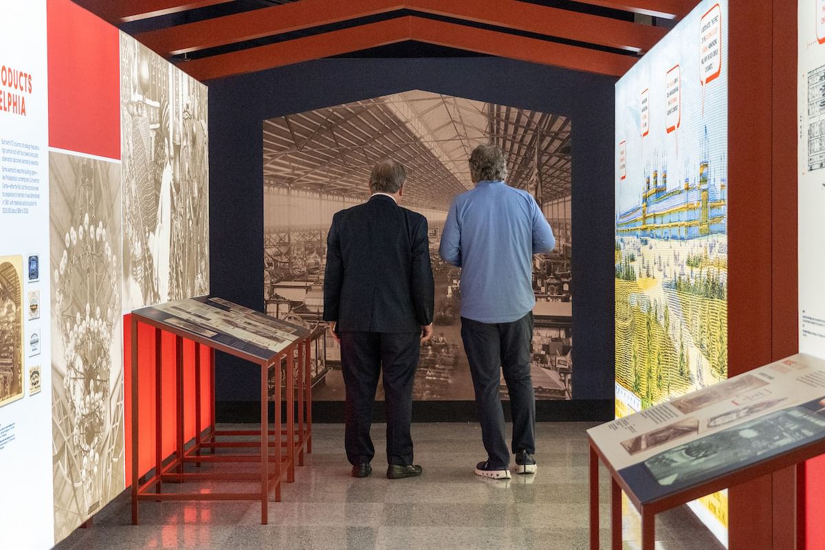 2 men seen from behind inside a colorful historic display, looking at a giant black & white photo of Philly's 1876 Centennial