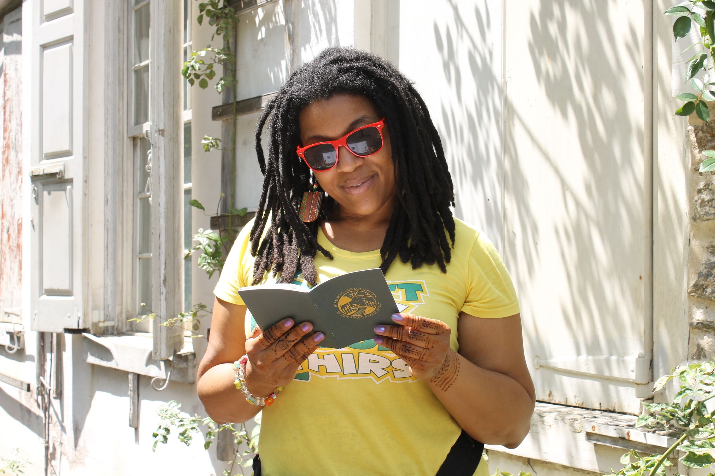A Black woman in sunglasses, locs, and a yellow shirt, outside on a sunny day, smiles at a small book open in her hands.