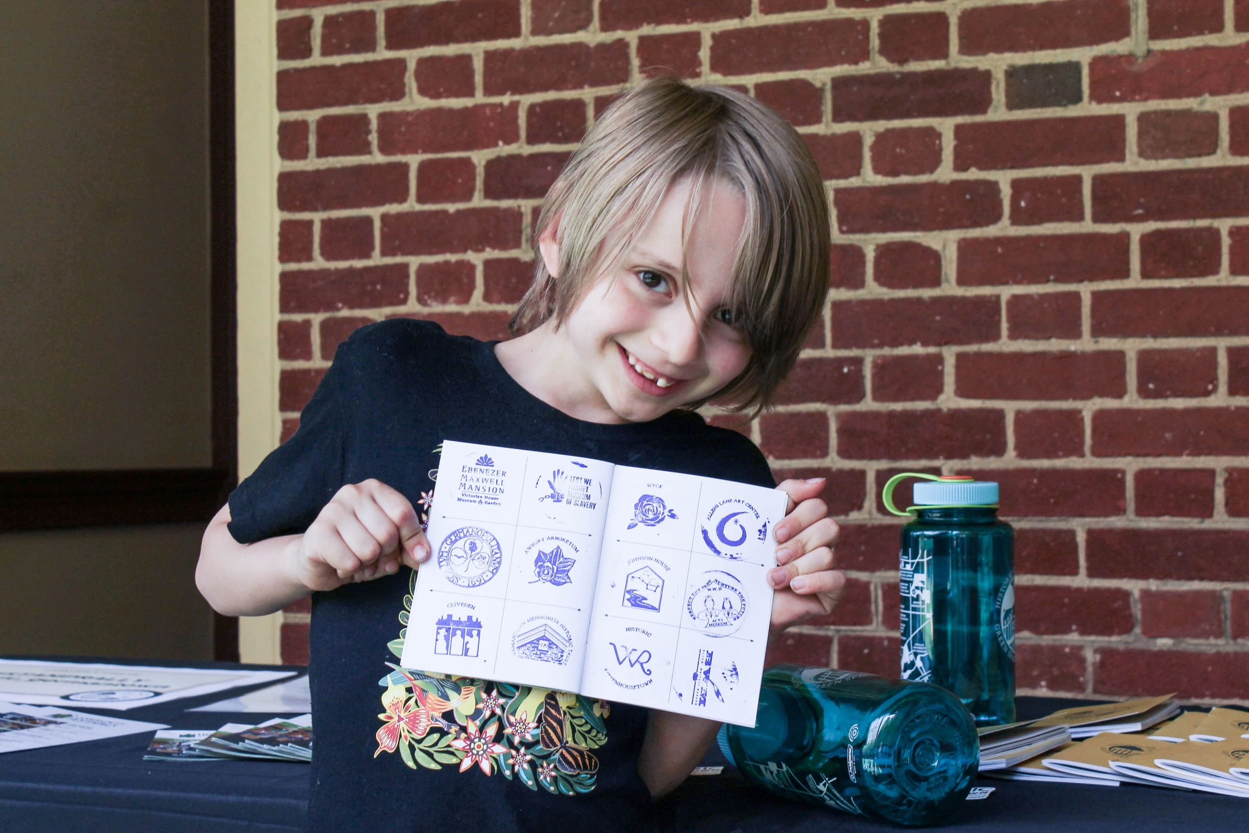 A smiling white boy proudly holds open a small book whose pages are stamped with different symbols in blue ink.