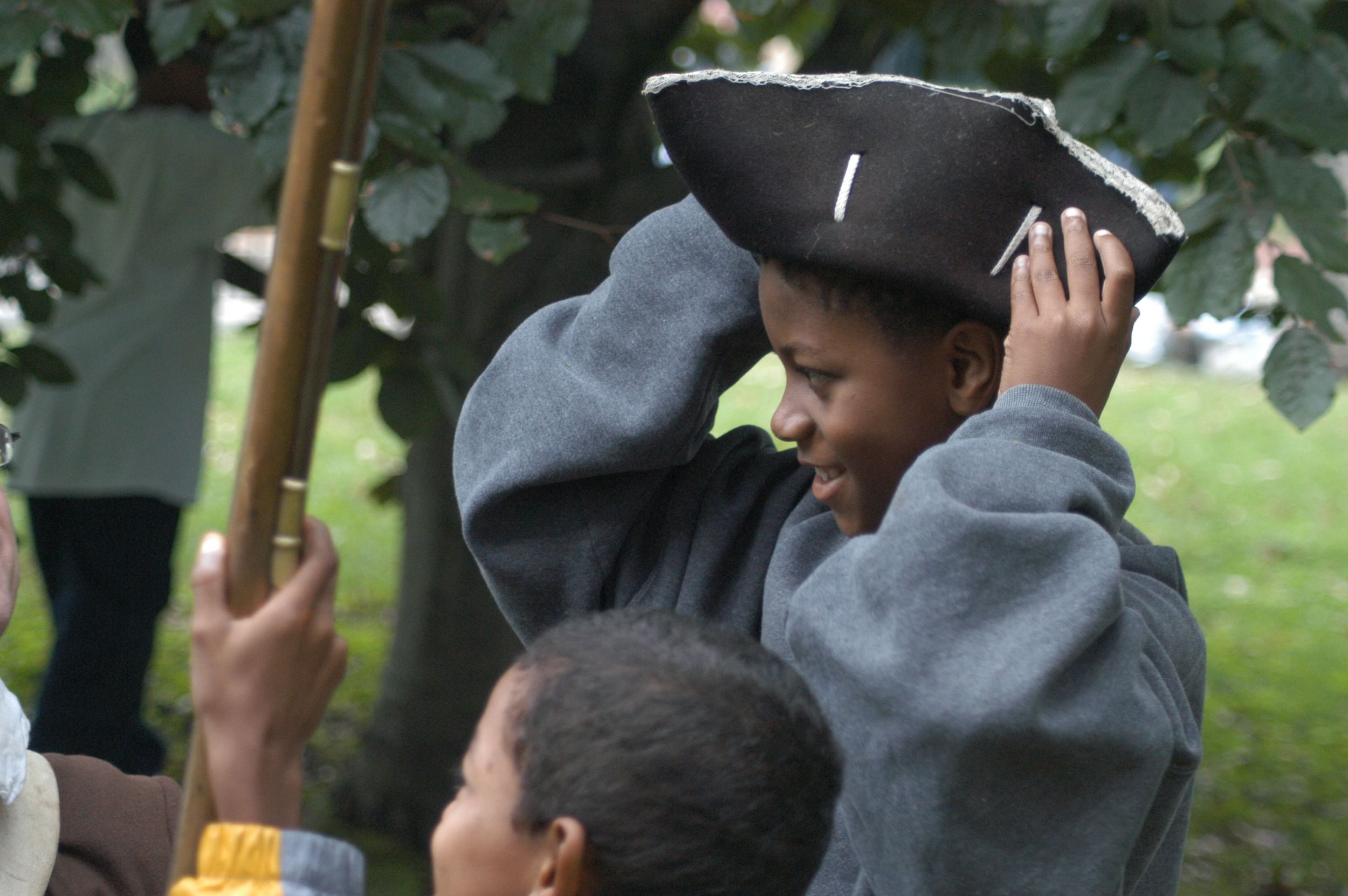 Closeup on a smiling Black boy in a gray sweatshirt, seen in profile trying on a tricorn hat.
