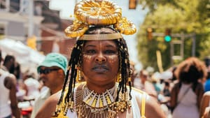 A woman dressed in a bright yellow dress with see-through sleeves, layers of necklaces, hair beads, and headdress