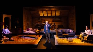 In a cozy, warmly lit cabin living room, Strathairn stands at center and the others sit or lie on couches and chairs.