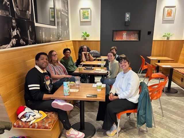 A multiracial group in casual clothes pose together smiling in the corner of a bright cafe.