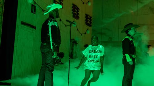 Gabrielle Hamilton, a Black woman in a sequined tee that reads "dream baby dream," leaps through green mist between 2 actors