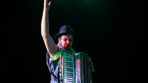 Harrold, in a black boulder & vest, throws one arm and leg in the air while playing an accordion in a wash of green light.