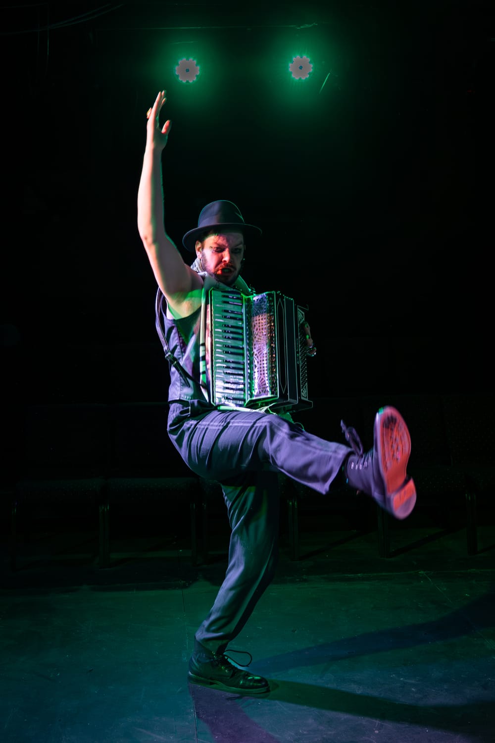 Harrold, in a black boulder & vest, throws one arm and leg in the air while playing an accordion in a wash of green light.