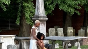 In a 17th-century graveyard, a Black woman with a blond buzz-cut caresses a younger Black woman sitting in a chair.