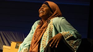 Muhammad, a young Black Muslim woman in brown hijab and light-blue tunic with brown embroidered leaves, laughs joyfully.