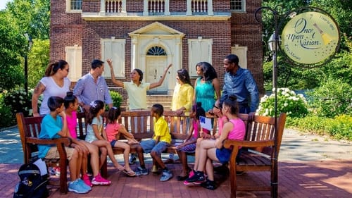 In front of a brick Colonial building, 13 people of different ages sit on a bench listening to a storyteller with raised arms