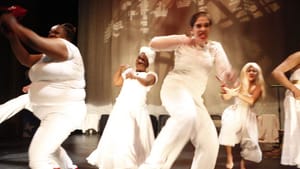 A group of five women in all white, expressive, thrash about on stage