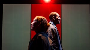 Yorke, a white woman, and DeShields, a Black man, stand in profile back-to-back, with a red light flaring overhead.