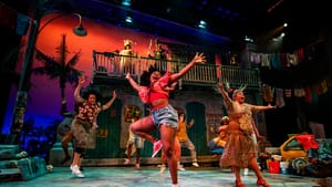8 actors dance exuberantly on a two-tier set with a junkyard and palm tree, with a rich orange and blue backdrop.