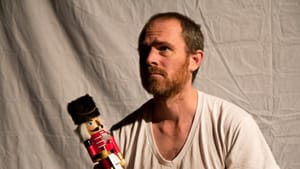 Davis, with short, gingery hair & beard, wearing a white tee, holds a wooden nutcracker doll and has a thoughtful expression.