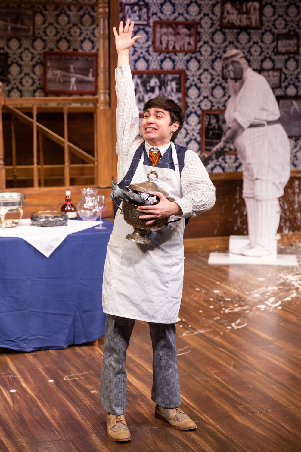 Gleason, in white apron, stands grinning with one hand raised as if in a classroom. He clutches a metal pot full of fake fish