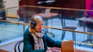 A woman sits at a table in a cafe wearing headphones, coat, and scarf. She arranges coffee beans spread out on the table