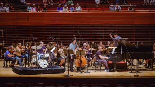 Stage-level photo of the casually dressed conductor and musicians rehearsing onstage while some people watch in seats above